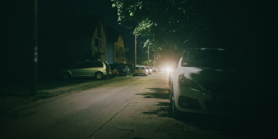 A white car parked on a dark street, slightly hidden by a tree.